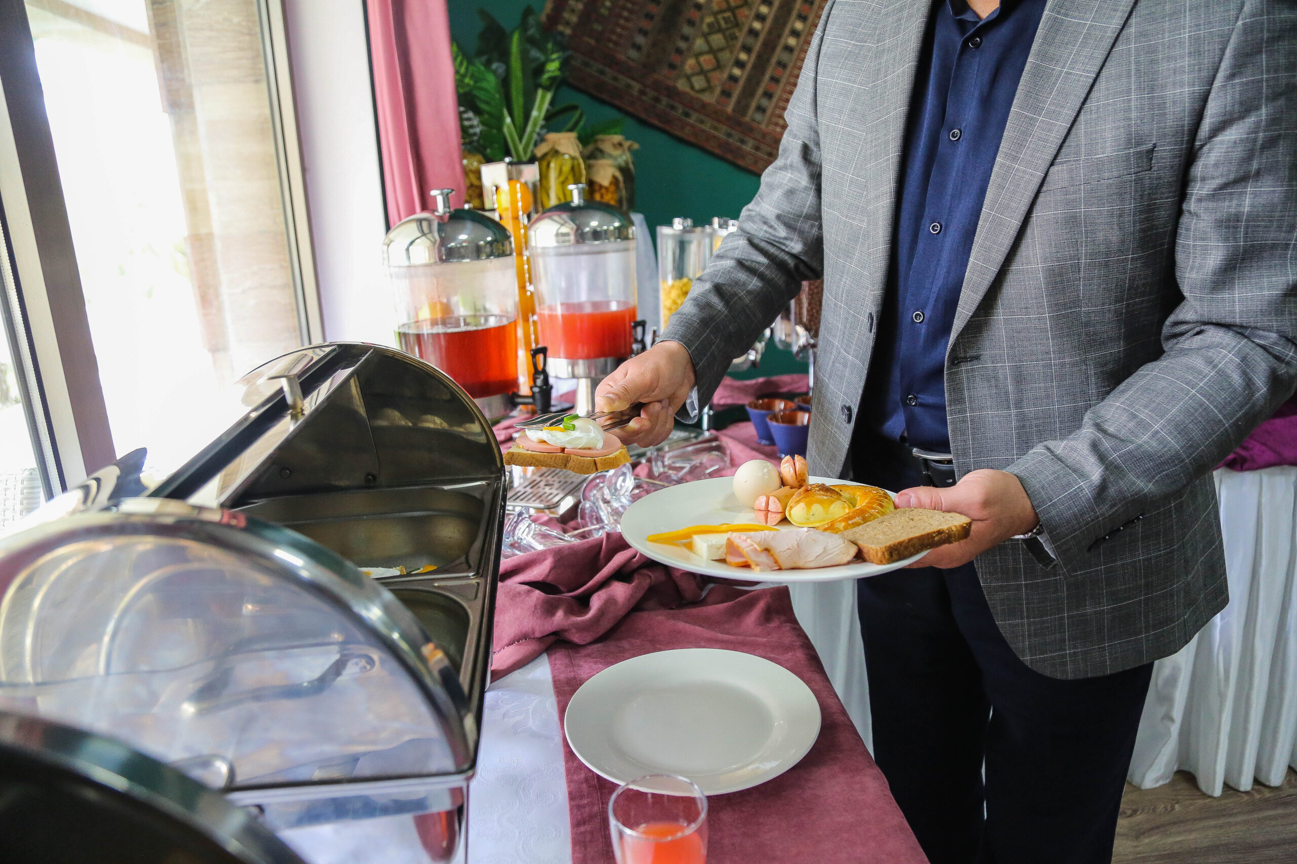 side view man lays breakfast food egg sausages bun toast and cheese on a plate from the open buffet